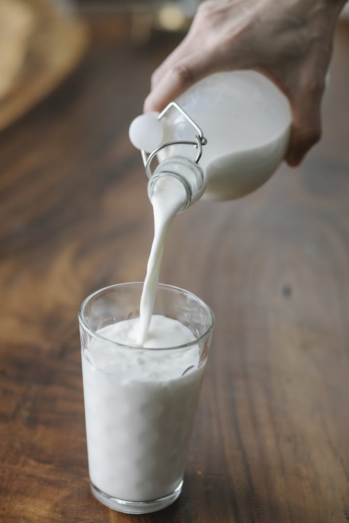 Crop anonymous person with bottle pouring milk into ornamental transparent glass on wooden table at home